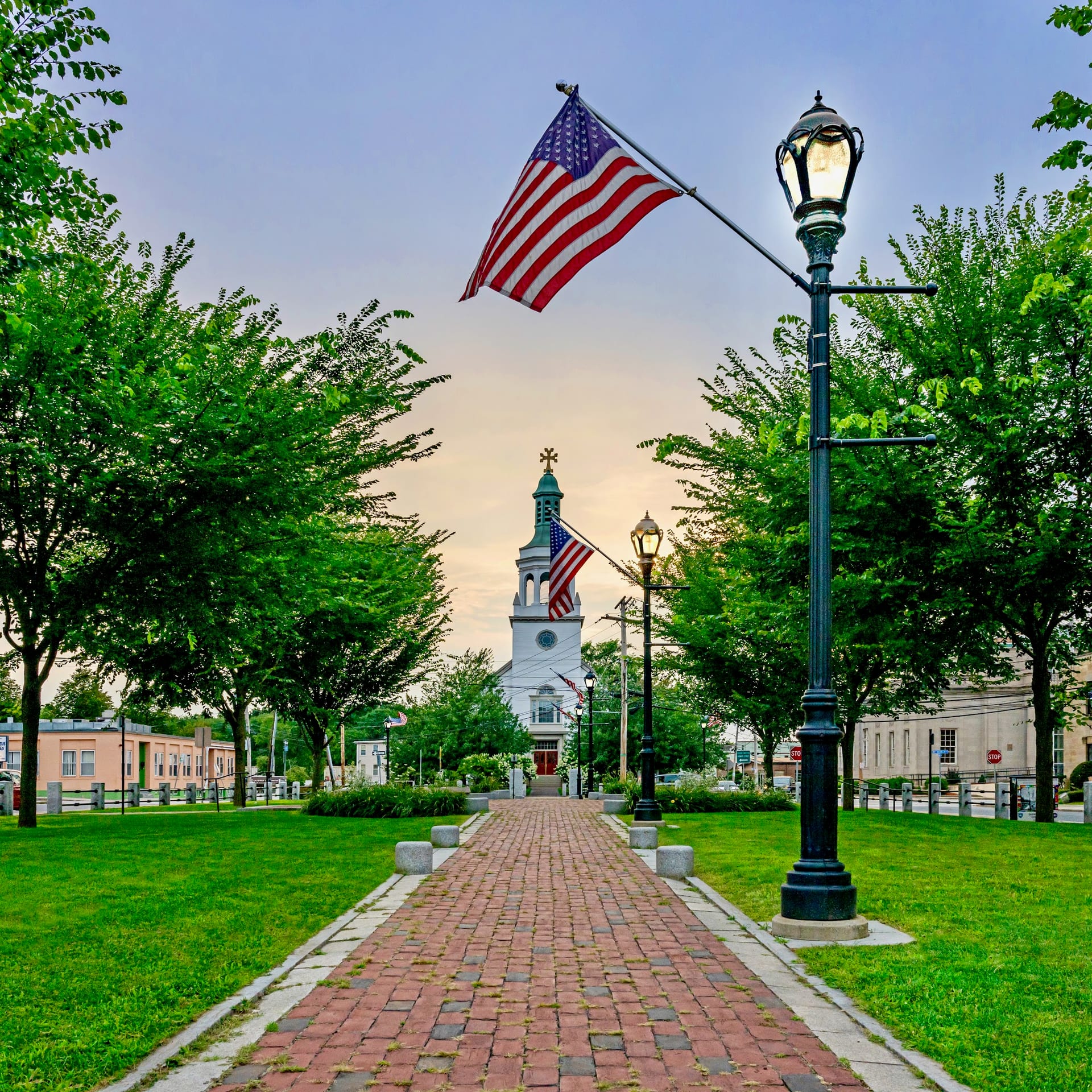 Tree-lined brick path leading to white church steeple with American flags
